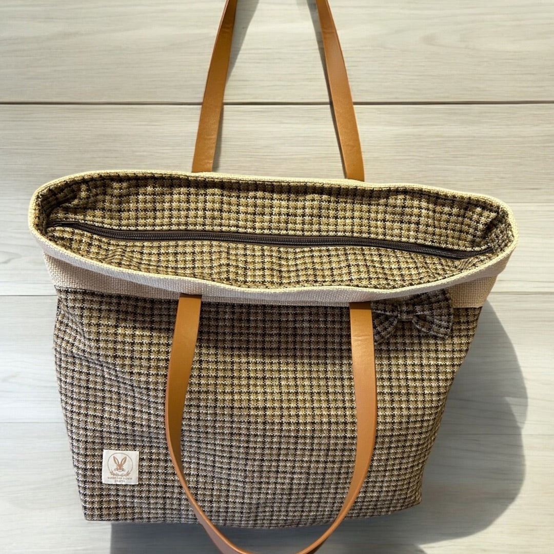 Tweed tote bag with leather handles, zip closure, and a decorative bow, standing upright.