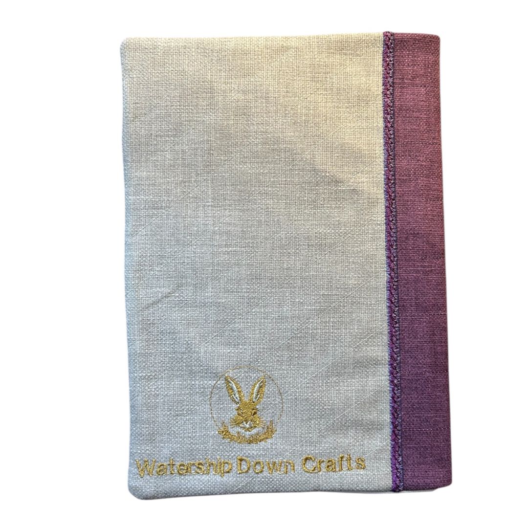 Back view of a handmade notebook cover in natural fabric with a purple spine, embroidered with the Watership Down Crafts logo in gold thread.