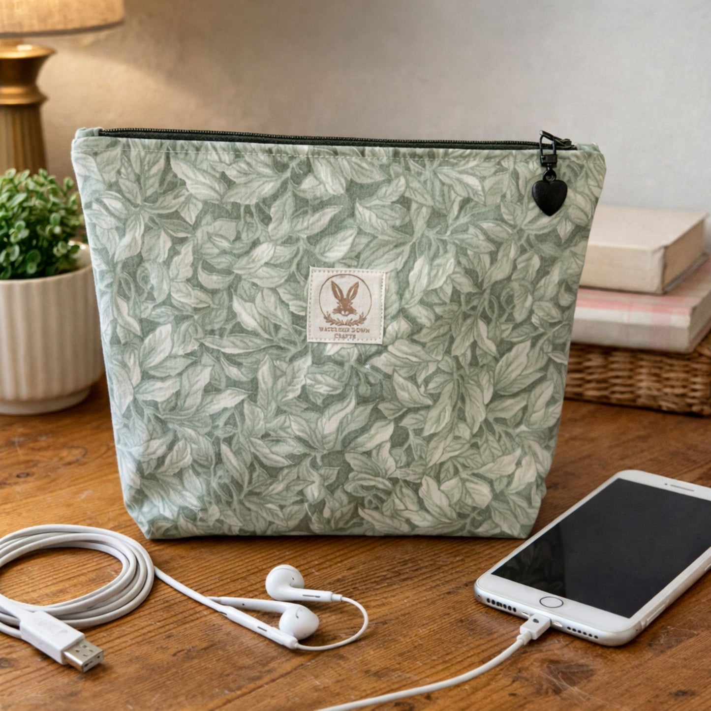 Green leaf-patterned pouch with a brand label on a wooden surface with a phone, earbuds, and cable.