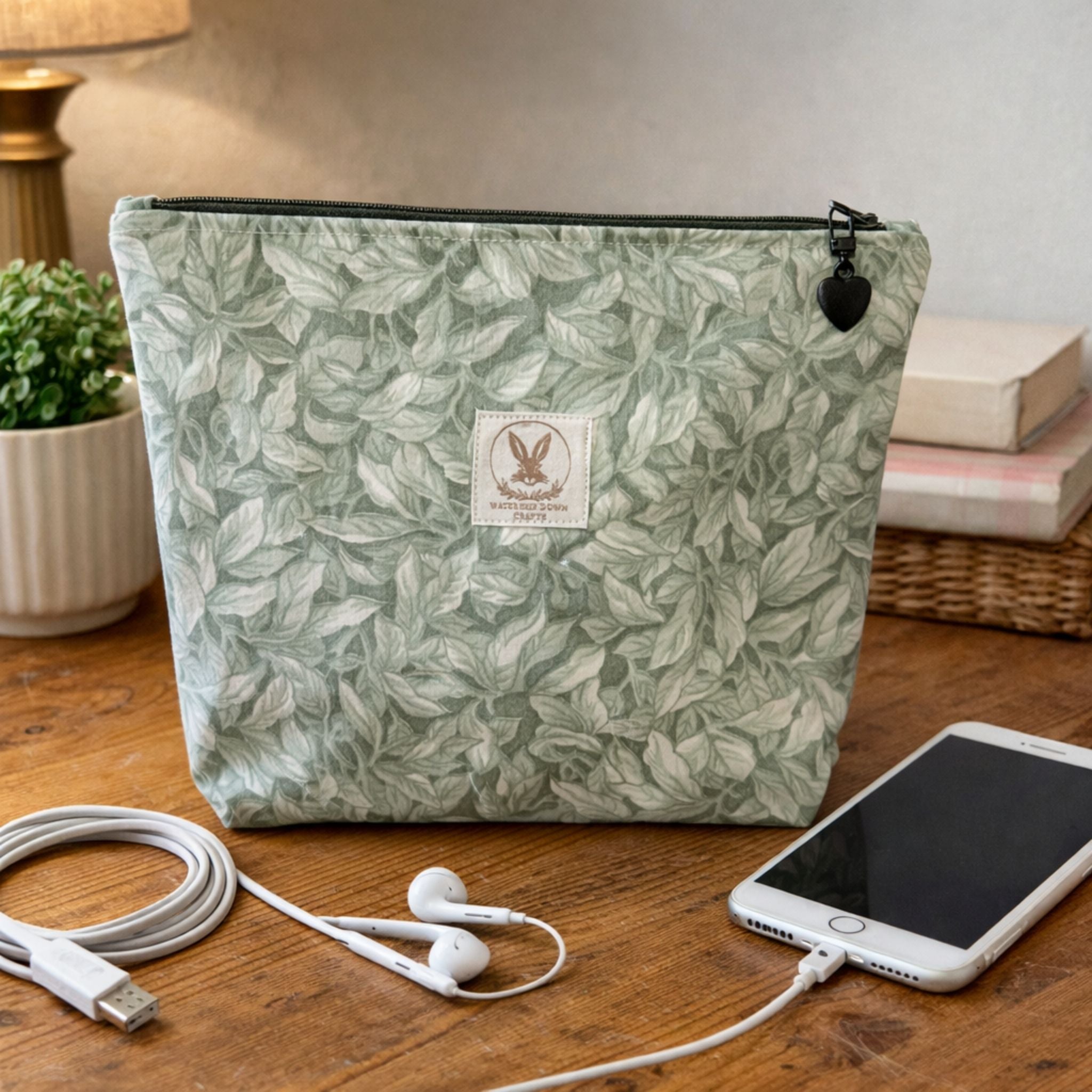 Green leaf-patterned pouch with a brand label on a wooden surface with a phone, earbuds, and cable.