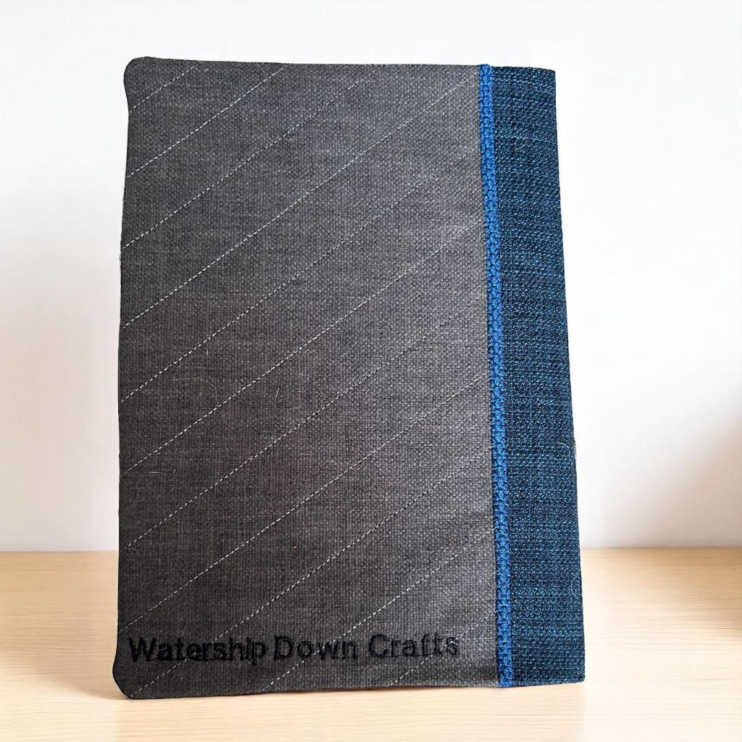 Back view of an embroidered A5 notebook cover in dark grey fabric with subtle diagonal stitching and a blue fabric spine.