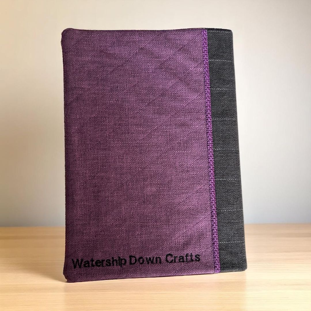 Back view of a purple and charcoal fabric A5 notebook cover with diagonal quilting and “Watership Down Crafts” embroidered in black.