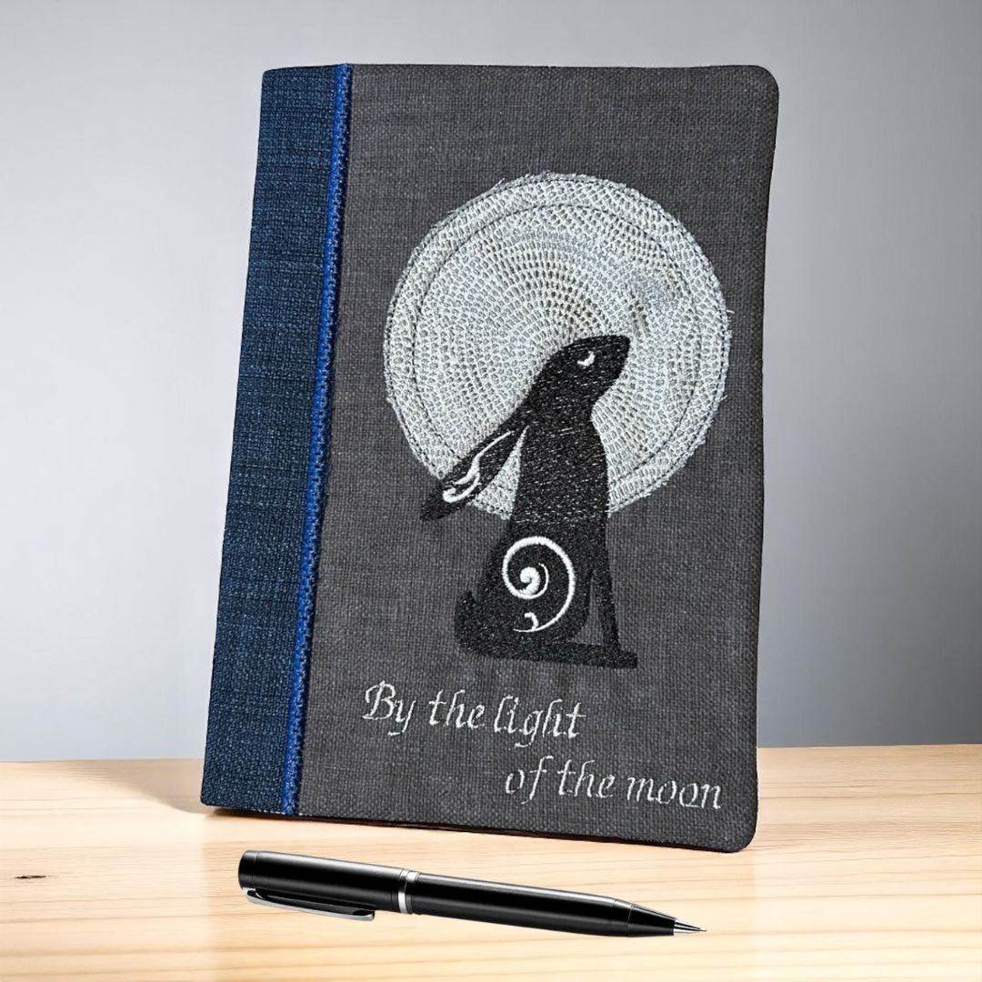 Embroidered A5 notebook cover featuring a black moon gazing hare against a full moon, with the words “By the light of the moon” stitched below. A black pen is placed in front.