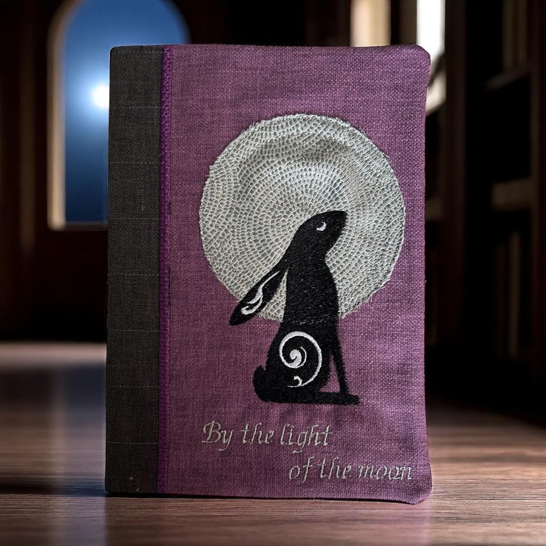 Purple embroidered A5 notebook cover with a moon gazing hare design and the words “By the light of the moon,” photographed in low light with a glowing background.
