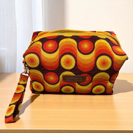 Handmade makeup bag with bold retro swirl fabric in orange, yellow, and brown tones, featuring a yellow zip and wrist strap. Made by Watership Down Crafts.