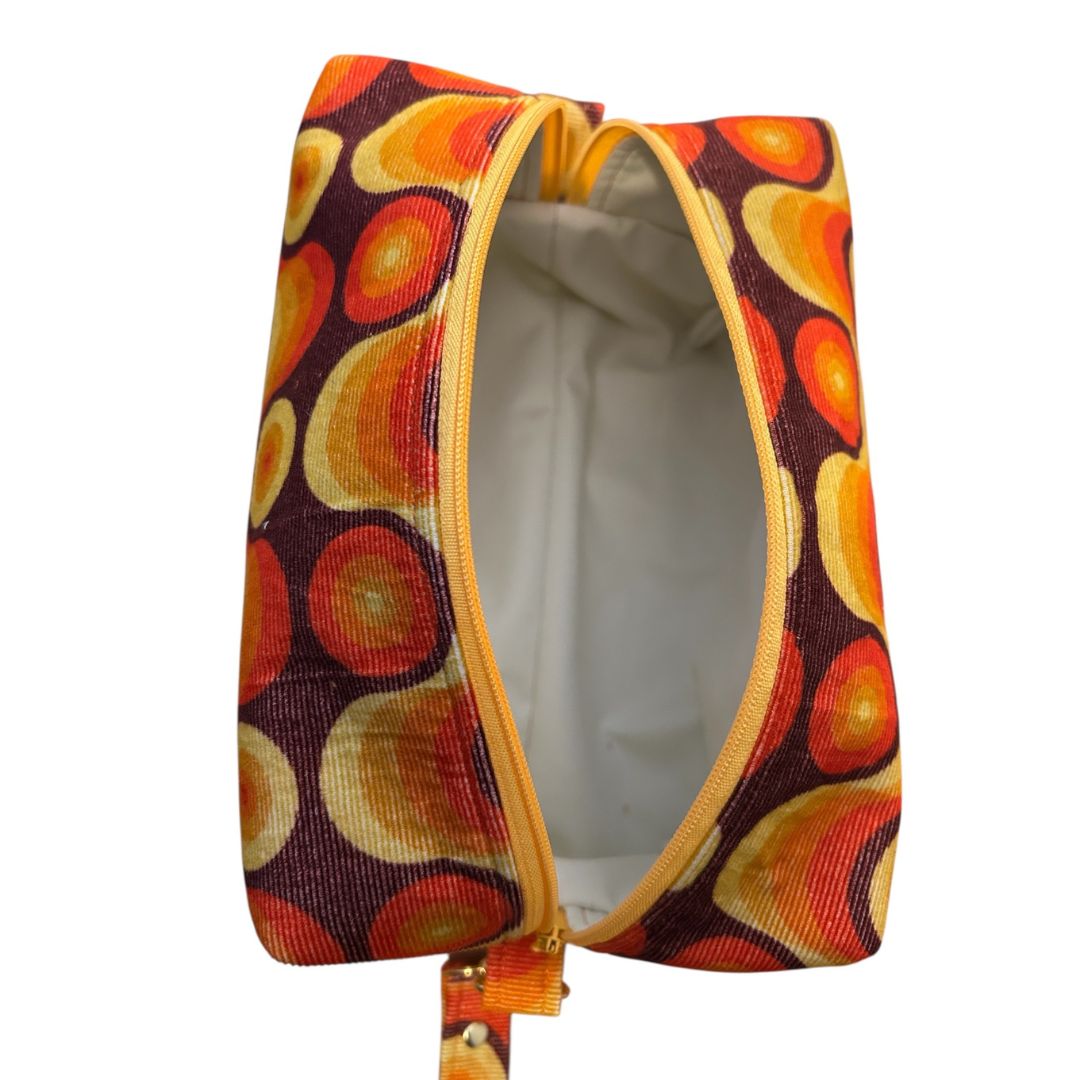 Top-down view of open retro makeup bag showing cream water-resistant lining, yellow zip, and bold swirl fabric in orange, yellow, and brown.