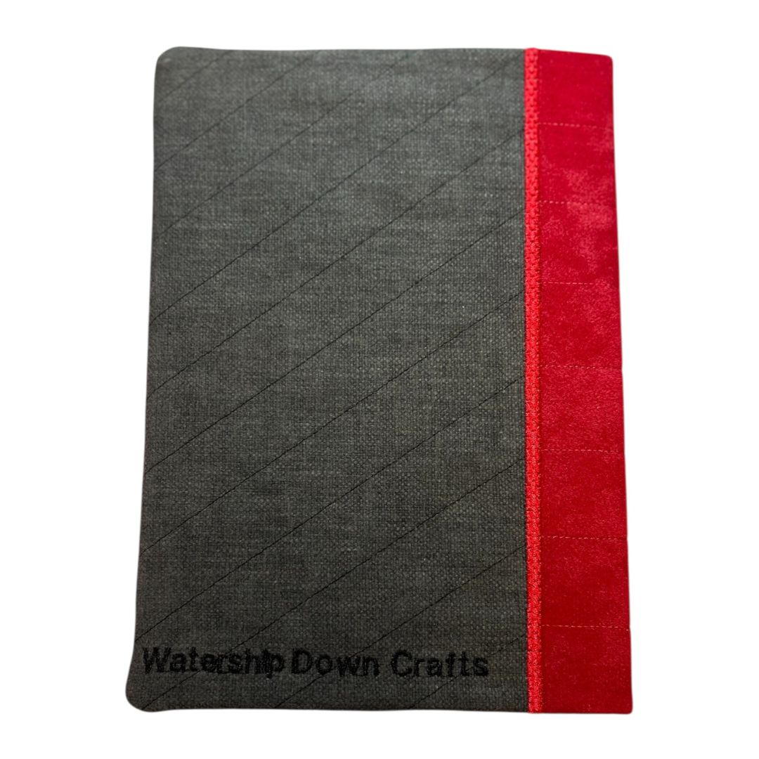 Back view of a handmade notebook cover in dark grey fabric with diagonal quilting, featuring a bold red fabric spine and black Watership Down Crafts logo embroidery.
