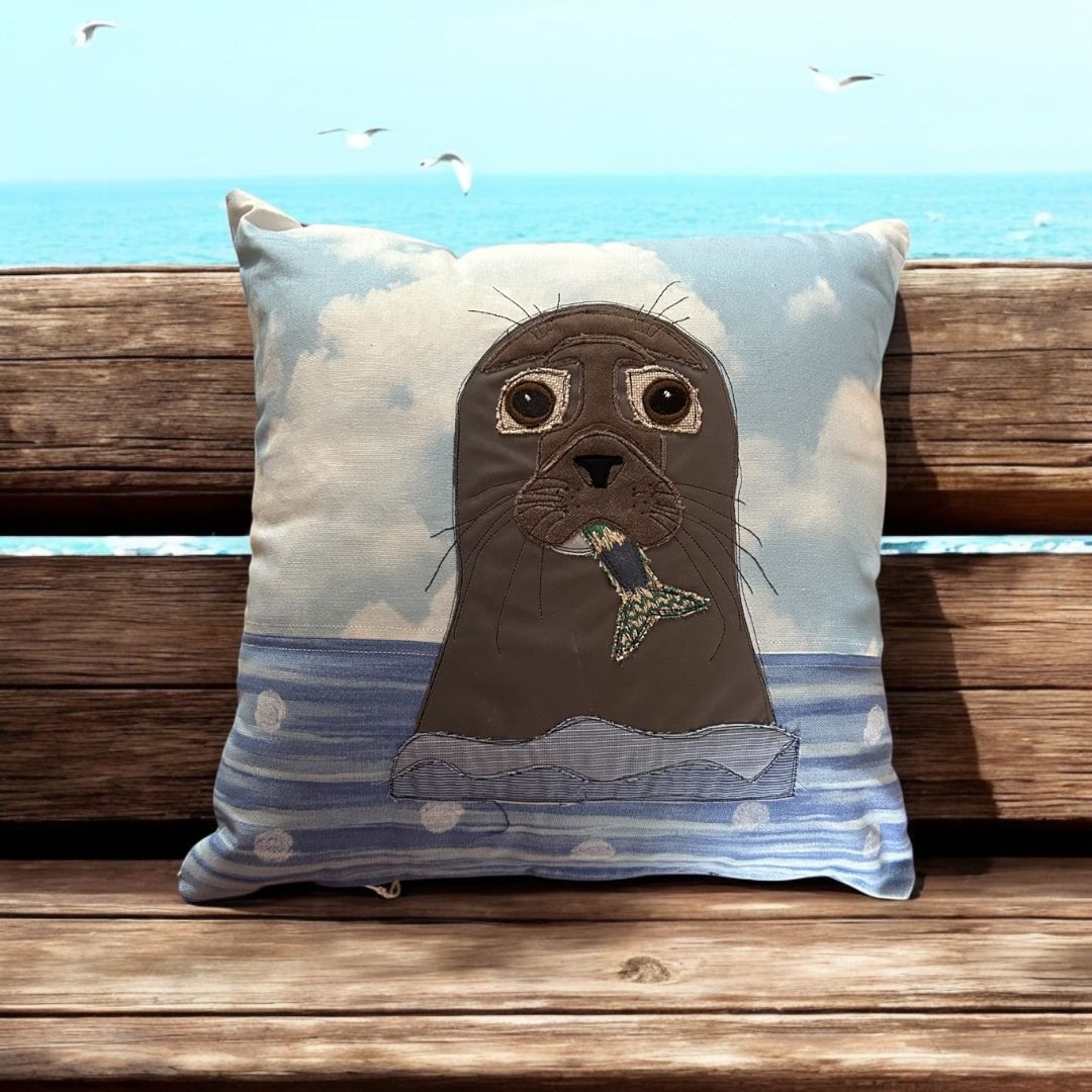 Raw-edge appliqué cushion featuring a seal holding a fish, set against a blue sky and ocean backdrop, photographed on a wooden bench by the sea with birds flying in the background.