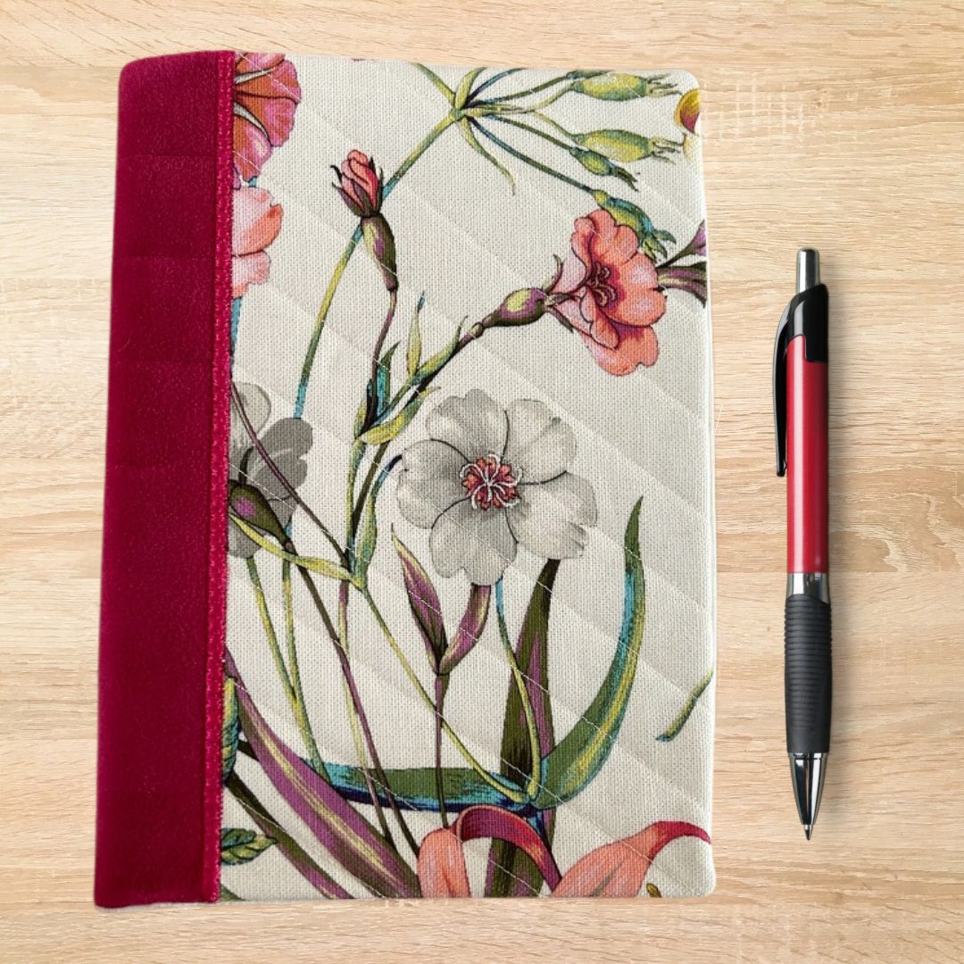 Front view of an A5 notebook cover featuring a colourful floral design on cream fabric with a deep red fabric spine, shown beside a red and black pen on a wooden surface.