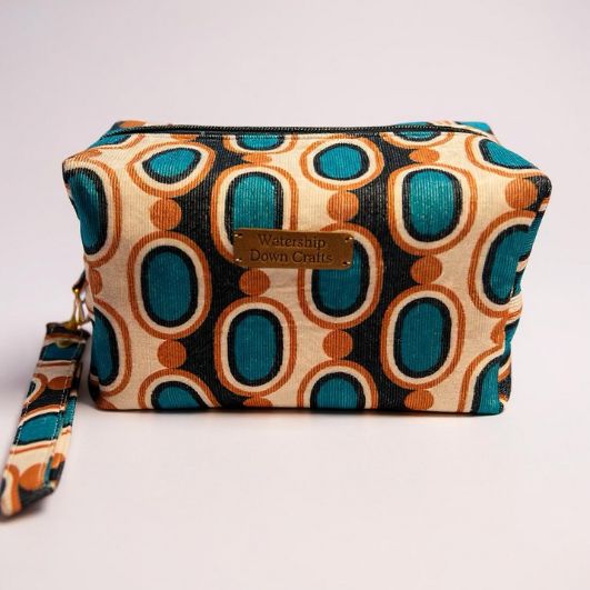 Front view of a boxed makeup pouch in bold retro blue, tan, and cream oval print with a detachable wrist strap and a Watership Down Crafts label.